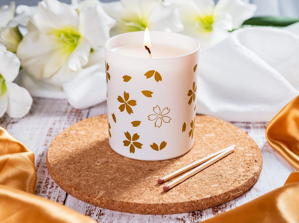 White candle with gold floral patterns on a cork coaster with white flowers in the background