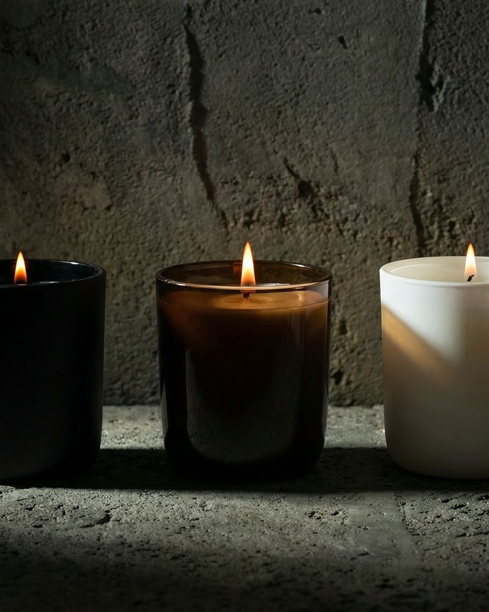 Three candles of different colors (black, brown, white) lit against a dark stone wall.